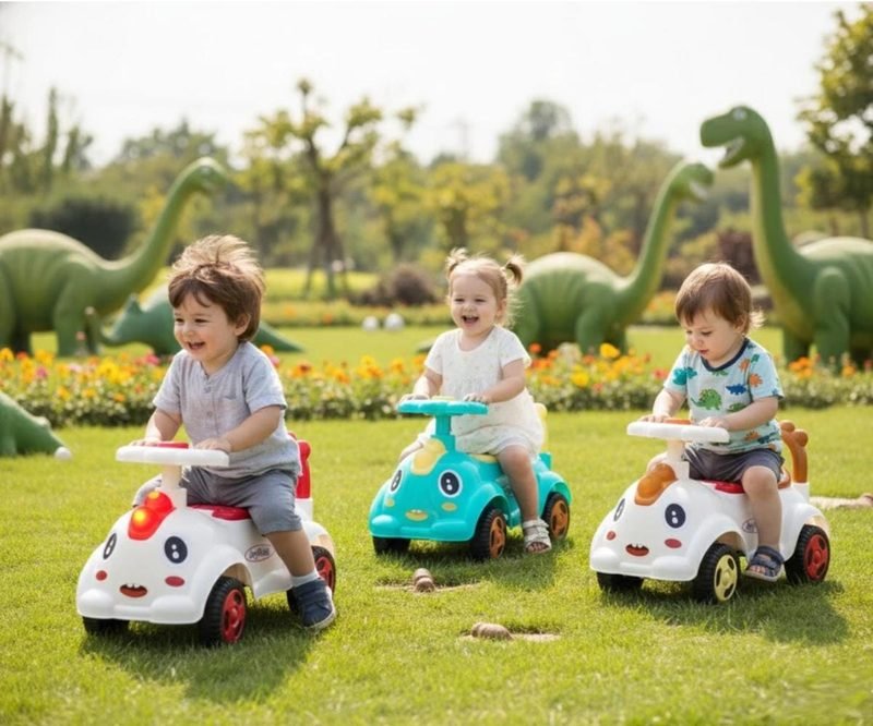Three toddlers riding Funfizz cartoon-face ride-on cars across the grass of a dinosaur-themed park